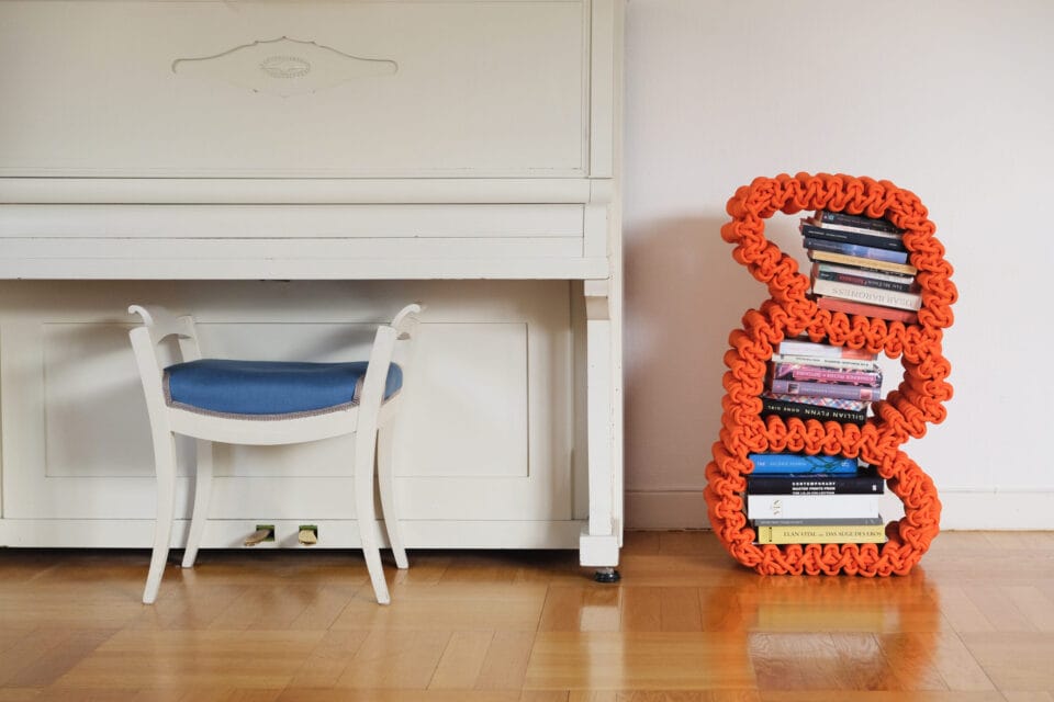 an orange, knitted nylon rope shelf with books on it, shown next to a white piano