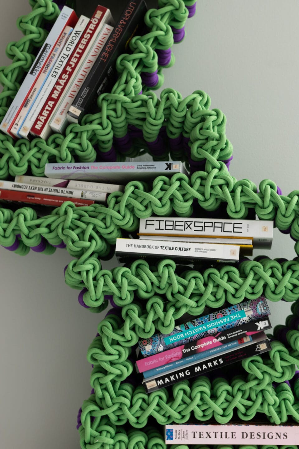 a detail of a green shelf made from thick nylon rope, with books in its openings