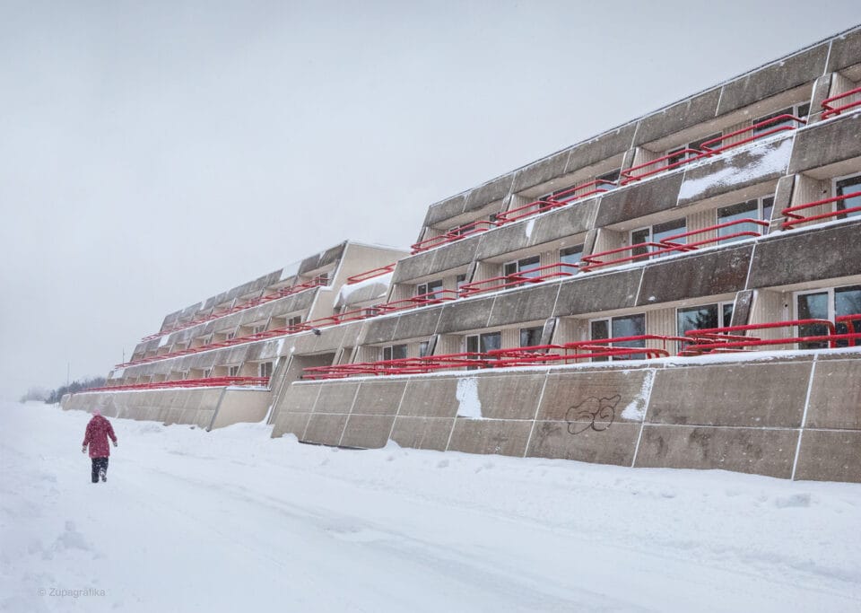 a figure in a red coat walks alongside a brutalist apartment block in an otherwise barren, snowy landscape