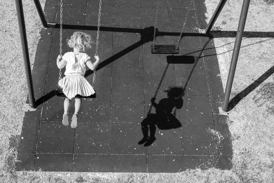 a black and white image of a child swinging with her shadow seeming to swing on the ground below