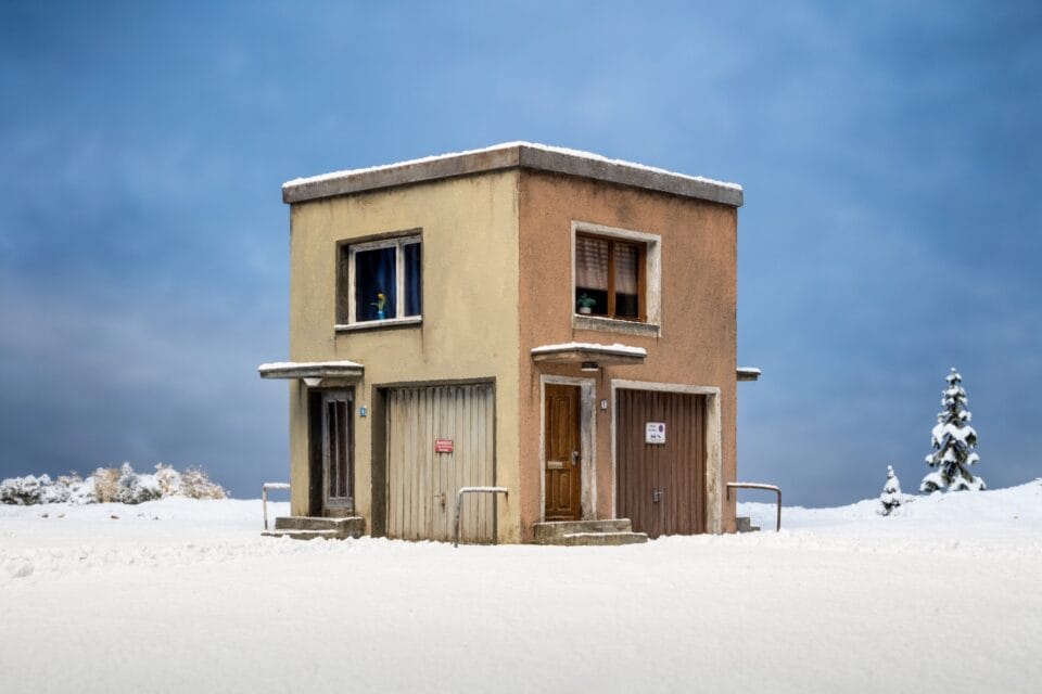 a photo of an uncanny miniature set showing a square building in an empty snow-covered landscape, with doors, windows, and garage doors on all sides