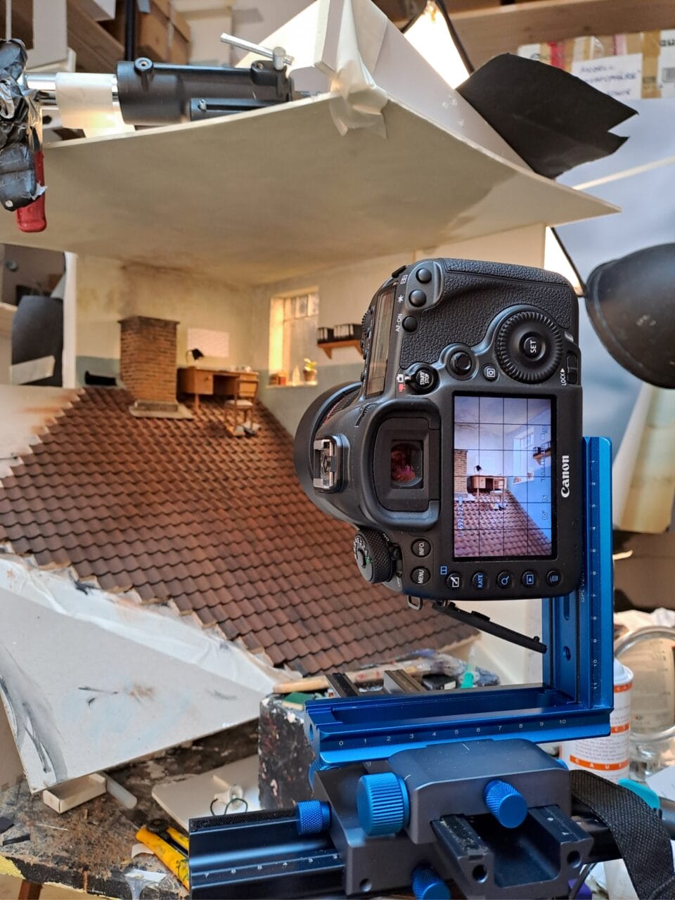 a behind-the-scenes photo of a miniature set with a desk inside of a room, on a steep tile roof, with a camera in the foreground showing the composition on its screen