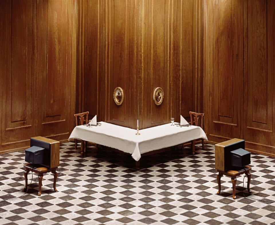 a photo of a miniature set showing a corner of a room with an L-shaped table, each with its own place setting and television