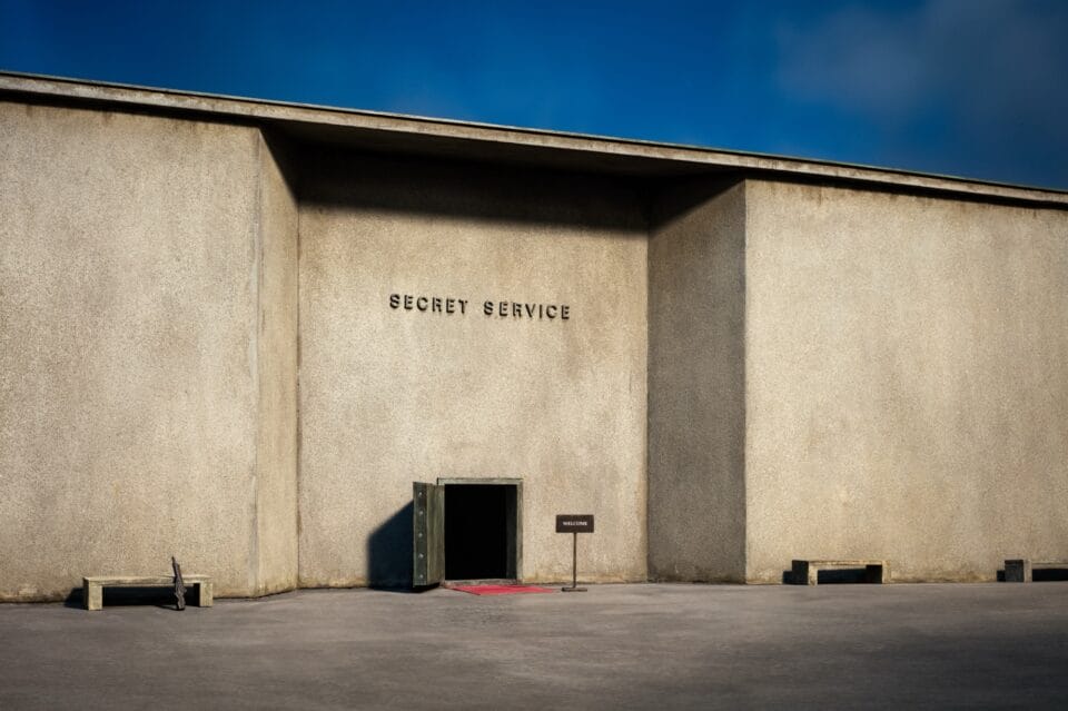 a photo of a miniature set sharing a brutalist building that reads "secret service" on top, with a wide-open door and a "welcome" sign outside